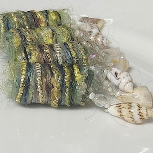 May include: Close-up of handmade jewelry components. The design features a combination of green, gold, and blue woven elements, accented with small seashells and clear beads. The components are arranged on a white surface.