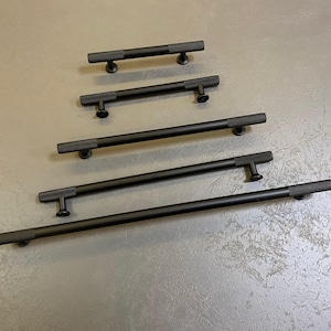 以下が含まれることがあります： さまざまな長さの黒いキャビネットプル。各プルは、テクスチャード加工されたダイヤモンドパターンのグリップと円筒形のバーを備えています。プルは灰色の表面に取り付けられており、洗練されたモダンなデザインを示しています。