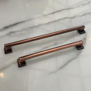 Könnte beinhalten: Zwei gebürstete Kupfer-Schrankgriffe mit rechteckiger Form und leicht abgerundeter Form. Die Griffe haben eine glatte, satinierte Oberfläche.