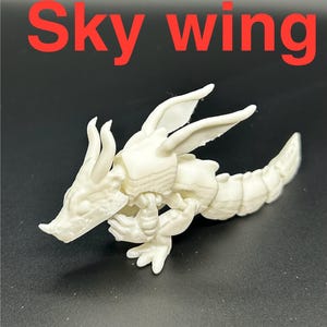 Op de afbeelding: Een witte, gelede drakenfiguur met de tekst "Sky wing" in het rood. De draak heeft gedetailleerde vleugels, een lange staart en staat op een donkere ondergrond. De figuur is waarschijnlijk 3D-geprint.