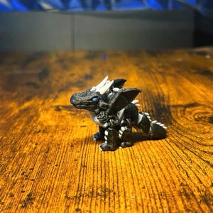 Pu&ograve; includere: Una piccola statuetta di drago articolata con un corpo nero testurizzato e accenti bianchi su corna, artigli e spine dorsali. La creatura ha occhi blu brillante e un design segmentato dettagliato che consente il movimento degli arti e della coda.