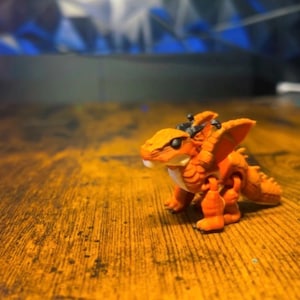 Pu&ograve; includere: Una piccola statuetta di drago arancione articolata con un corpo testurizzato e squamato, occhi neri e piccole ali. La creatura &egrave; raffigurata in posizione eretta con giunture visibili su spalle e fianchi, caratterizzata da un design modellato.