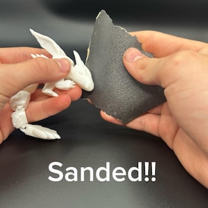 Op de afbeelding: Een witte, gelede drakenfiguur wordt geschuurd met schuurpapier. De draak heeft vleugels en een lange staart. De tekst "Sanded!!" staat in het wit onderaan de afbeelding.