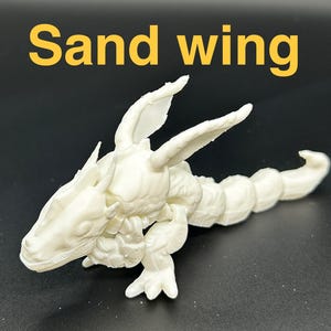 Op de afbeelding: Een cr&egrave;mekleurig, gearticuleerd drakenbeeldje met de tekst "Sand wing" in het geel. De draak heeft een gedetailleerde kop, vleugels en een gesegmenteerd lichaam. Het beeldje staat tegen een donkere achtergrond.