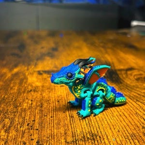Pu&ograve; includere: Una piccola statuetta di drago articolata con un corpo blu e verde acqua vibrante con punti gialli luminosi. La creatura presenta corna nere, ali bordate di arancione e una superficie dettagliata e strutturata, vista di profilo.