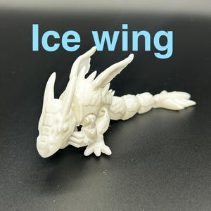 Op de afbeelding: Een witte 3D-geprinte drakenfiguur met de tekst "Ice wing" in blauw. De draak heeft gedetailleerde vleugels, een gesegmenteerd lichaam en een gedetailleerde kop. De figuur staat op een donkere ondergrond.