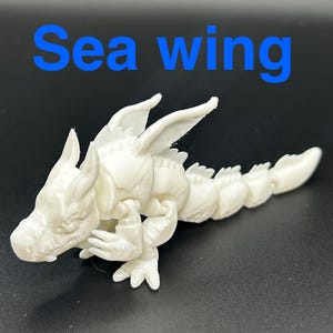 Op de afbeelding: Een witte, beweeglijke drakenfiguur met de tekst "Sea wing" in het blauw. De draak heeft een gedetailleerde kop, vleugels en een gesegmenteerd lichaam. De figuur staat op een donkere ondergrond.