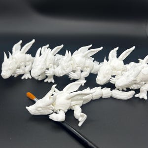 Op de afbeelding: Verschillende witte, beweegbare drakenfiguren zijn gerangschikt op een donkere ondergrond. De draken hebben gedetailleerde vleugels, poten en staarten. Een penseel met een oranje kop ligt op een van de draken, wat suggereert dat ze worden beschilderd.