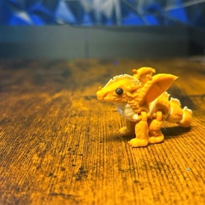 Pu&ograve; includere: Una piccola statuetta di drago articolata di un vivace colore giallo dorato. La creatura presenta un corpo squamato e testurizzato, ali prominenti, una testa definita con una piccola cresta e una lunga coda. Poggia su quattro zampe con giunture.