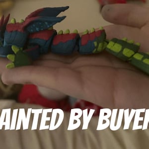 Op de afbeelding: Een kleurrijk, gearticuleerd drakenbeeldje in tinten blauw, rood en groen. De draak heeft vleugels en een gesegmenteerd lichaam. De tekst "PAINTED BY BUYER" wordt in witte, vette letters weergegeven.