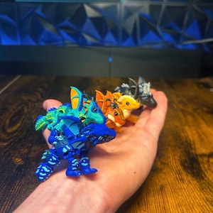 Pu&ograve; includere: Una collezione di quattro piccole statuette di drago articolate in vari colori, tra cui un drago blu brillante, uno turchese e giallo, uno arancione e uno nero e bianco, tutti caratterizzati da squame, ali e corna dettagliate in miniatura.