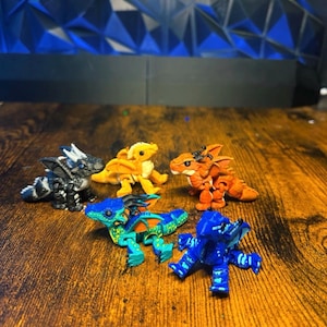 Pu&ograve; includere: Una collezione di cinque piccole statuette di drago articolate in vari colori, tra cui bianco e nero, giallo, arancione, verde acqua con accenti arancioni e blu solido. Ogni drago presenta squame, ali e corna dettagliate in miniatura.