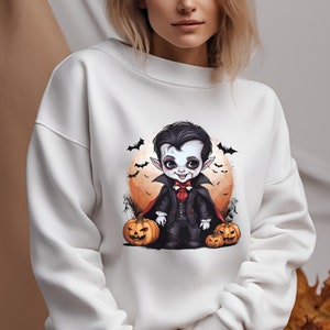 Könnte beinhalten: Weißes Sweatshirt mit einem Cartoon-Vampir-Design. Die Illustration zeigt einen Vampir in einem schwarzen Anzug, roter Fliege und Umhang, mit Kürbissen, Fledermäusen und einem orangefarbenen Mond, für Halloween.