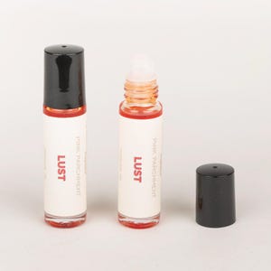 May include: Two small, clear glass perfume bottles with black caps and a white label. The label reads "LUST" in red, with "Pink Parchment" below. One bottle is open, revealing a rollerball applicator and pink liquid. The other bottle is closed.