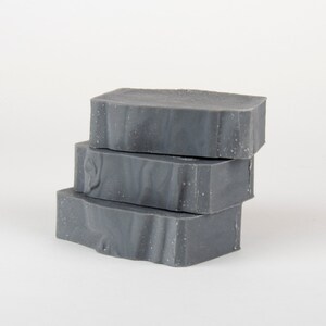 May include: Three stacked bars of gray soap with a textured surface. The soap has a marbled appearance with lighter gray swirls throughout. The edges of the soap are slightly uneven, giving it a handmade look.