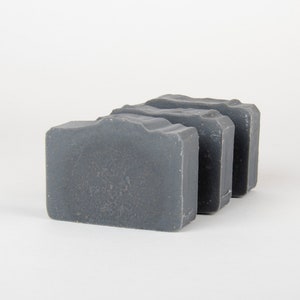 May include: Three rectangular bars of gray soap are arranged in a row. The soap has a textured surface and a slightly wavy top edge. The background is plain white.