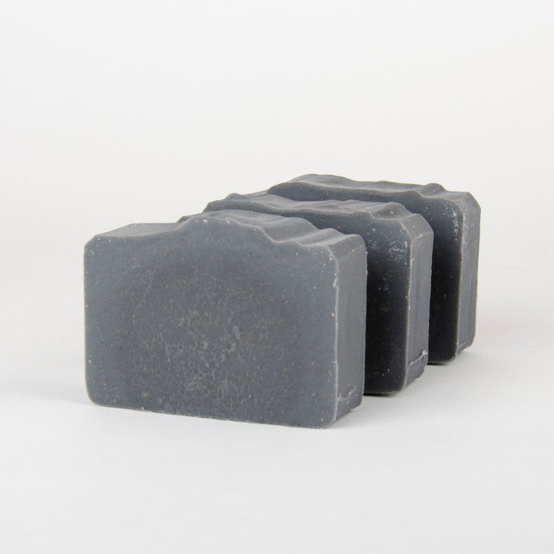 May include: Three rectangular bars of gray soap are arranged in a row. The soap has a textured surface and a slightly wavy top edge. The background is plain white.