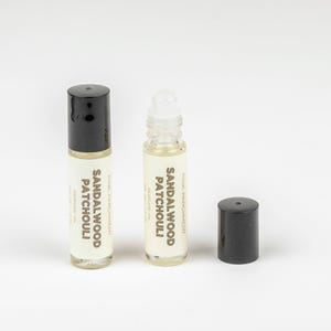 Peut inclure: Deux flacons d'huile parfumée en verre transparent avec des bouchons noirs et des étiquettes blanches. Les étiquettes indiquent "Sandalwood Patchouli" et "Pink Parchment". Un flacon est sans bouchon. L'huile parfumée est jaune clair.