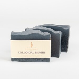 May include: Three dark gray bars of soap are arranged on a white background. The front bar has a cream-colored label with the text "COLLOIDAL SILVER" and "COLLOIDAL SILVER SOAP". The net weight is approximately 4.5 oz / 127 g.
