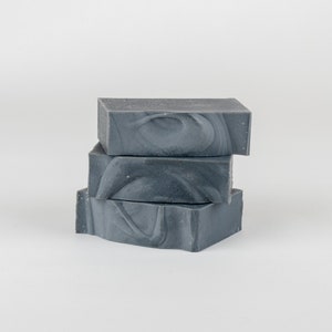 May include: Three stacked bars of gray soap with a marbled design. The soap bars are rectangular and have a textured appearance. The soap is a dark gray color with lighter gray swirls.