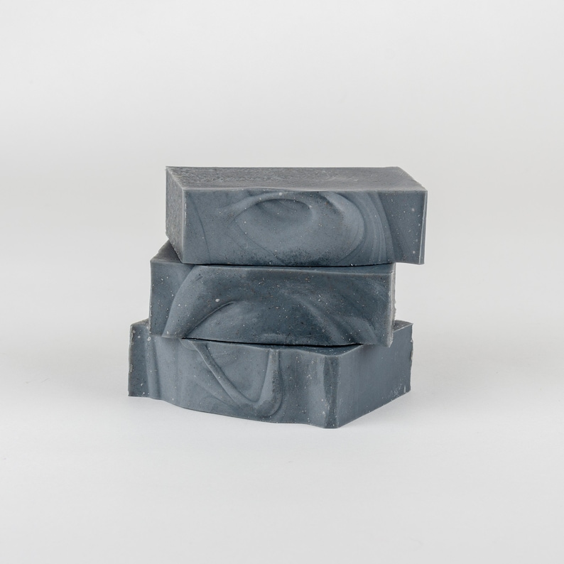 May include: Three stacked bars of gray soap with a marbled design. The soap bars are rectangular and have a textured appearance. The soap is a dark gray color with lighter gray swirls.