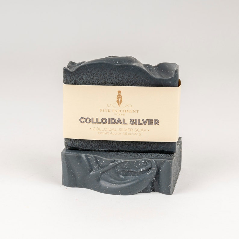 May include: Two bars of dark gray colloidal silver soap with a cream-colored label. The label reads "Pink Parchment Soaps" and "Colloidal Silver". The soap is approximately 4.5 oz/127 g. The soap has a textured surface and a decorative swirl design.