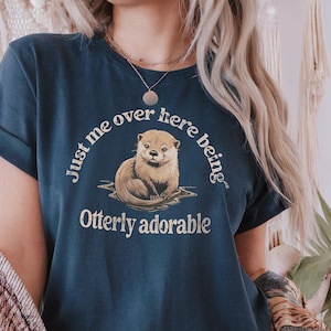 May include: A navy blue t-shirt with a graphic of an otter and the text "Just me over here being otter-ly adorable".