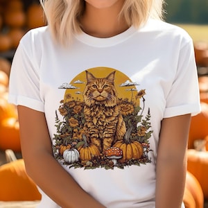 May include: White t-shirt with a graphic of a brown tabby cat sitting in a field of sunflowers and pumpkins. The cat is surrounded by fall foliage and mushrooms.