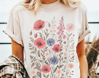 Può includere: T-shirt beige con stampa floreale di papaveri rosa, fiordalisi blu e altri fiori selvatici. L'illustrazione botanica include foglie e steli dettagliati. La maglietta ha le maniche arrotolate.