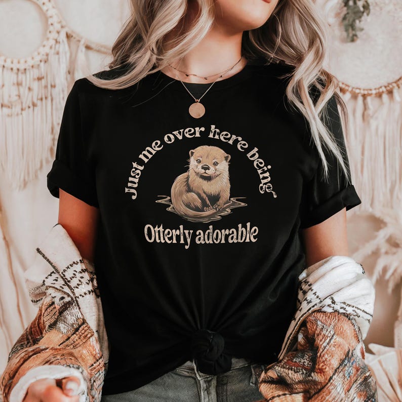 May include: Black t-shirt with a brown otter illustration and the text "Just me over here being Otterly adorable".
