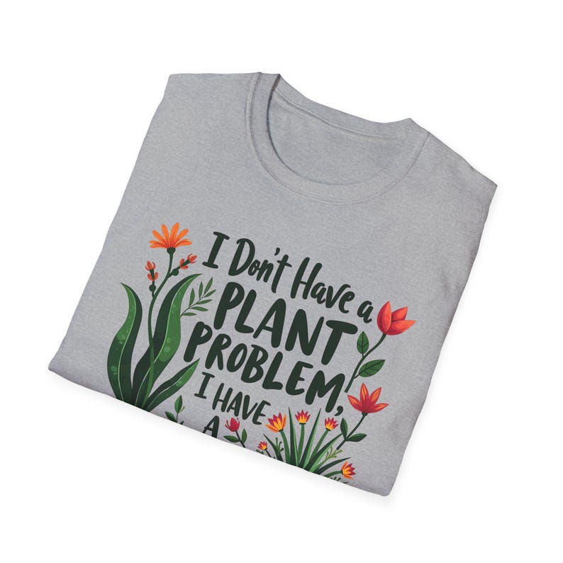 May include: A pale grey crew neck t-shirt featuring the text "I Don't Have a PLANT PROBLEM, I HAVE A" in green and white. The shirt is decorated with green leaves and orange and red flowers.