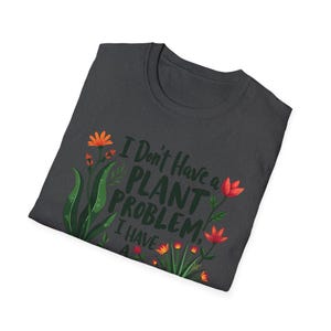 May include: Dark grey t-shirt featuring the text "I Don't Have a PLANT PROBLEM, I Have..." and floral illustrations in orange, red, and green. The shirt is folded, displaying the design on the front.