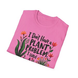 May include: A pink t-shirt featuring the text "I Don't Have a Plant Problem, I Have A..." and floral designs in orange, green, and yellow. The shirt is folded and presented on a white backdrop.