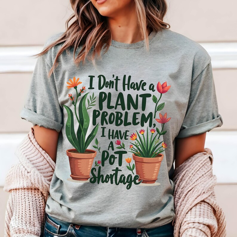 May include: A pale grey t-shirt with the text "I Don't Have a Plant Problem, I Have a Pot Shortage." The shirt features illustrations of potted plants with orange and pink flowers. The shirt has rolled-up sleeves.