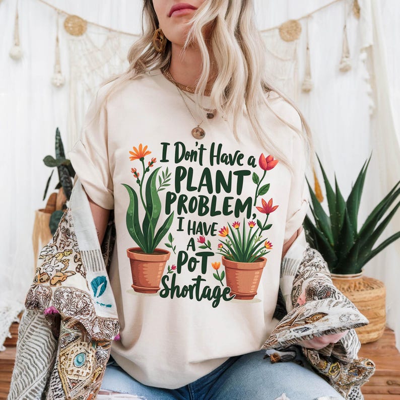 May include: A cream-coloured t-shirt with the slogan "I Don't Have a Plant Problem, I Have a Pot Shortage." The design includes illustrations of potted plants with orange and red flowers. The t-shirt is a relaxed, casual style.
