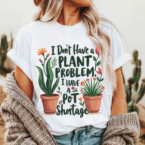 May include: White t-shirt with the text "I Don't Have a Plant Problem, I Have a Pot Shortage." The shirt features illustrations of potted plants with green leaves and orange and red flowers. The shirt is worn by a person.