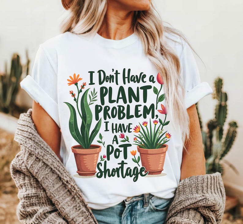 May include: White t-shirt with the text "I Don't Have a Plant Problem, I Have a Pot Shortage." The shirt features illustrations of potted plants with green leaves and orange and red flowers. The shirt is worn by a person.