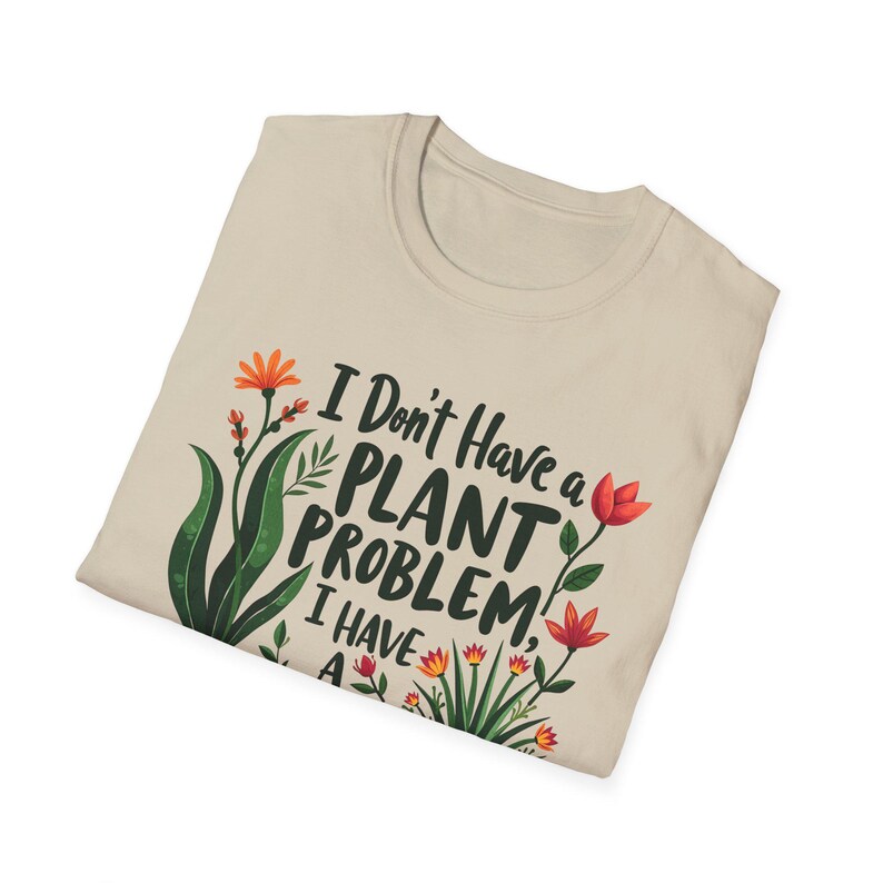 May include: A beige t-shirt featuring the text "I Don't Have a Plant Problem, I Have A..." and floral designs in orange, red, and green. The shirt is folded, displaying the front design. A casual, botanical-themed garment.