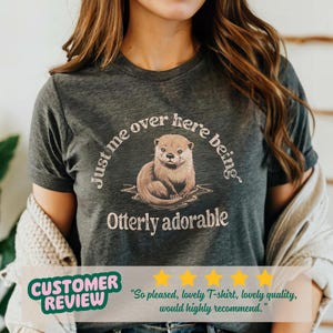 May include: A dark gray t-shirt with a graphic of an otter in water and the text "Just me over here being Otterly adorable".