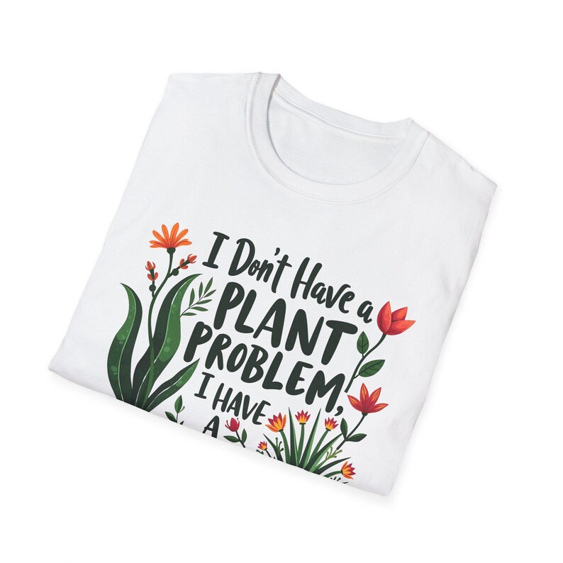 May include: White t-shirt featuring the text "I Don't Have a Plant Problem, I Have A..." with a floral design of green leaves and orange and red flowers. The shirt is folded.