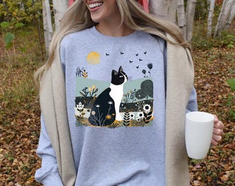 May include: A gray sweatshirt with a black and white cat illustration. The cat is sitting in a field of flowers and looking up at a flock of birds flying in the sky. The sun is shining in the background.
