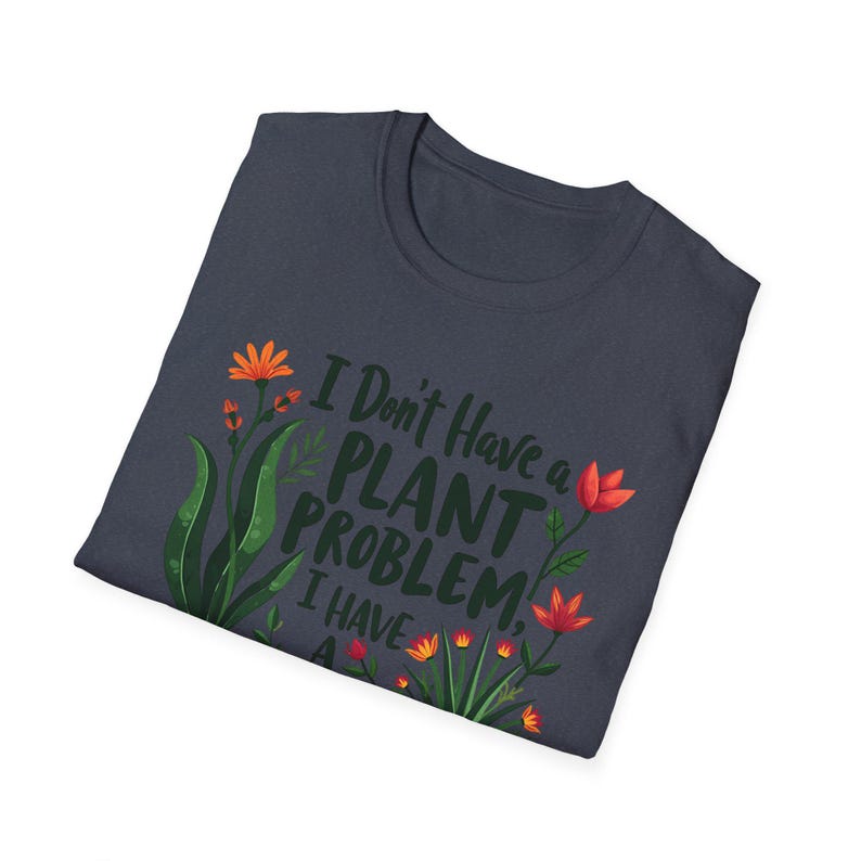May include: Dark navy blue t-shirt featuring the text "I Don't Have a Plant Problem, I Have A..." and floral illustrations in orange, red, and green. The shirt is folded, displaying the design.