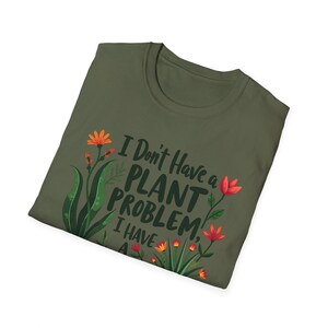 May include: Khaki green t-shirt featuring the text "I Don't Have a PLANT PROBLEM, I HAVE A..." and floral designs in orange and red. The shirt is folded and presented against a white backdrop.