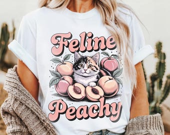 Könnte beinhalten: Weißes T-Shirt mit einer Grafik einer Katze und Pfirsichen. Der Text "Feline Peachy" steht in Retro-Schriftart über und unter dem Bild. Das Design zeigt eine Schildpattkatze und Pfirsiche in Rosa- und Pfirsichtönen.