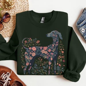 May include: Dark green sweatshirt with a floral dog design in pink, blue, and green. The dog is surrounded by floral and foliage elements. The sweatshirt has a crew neck and long sleeves. The design has a nature-inspired aesthetic.