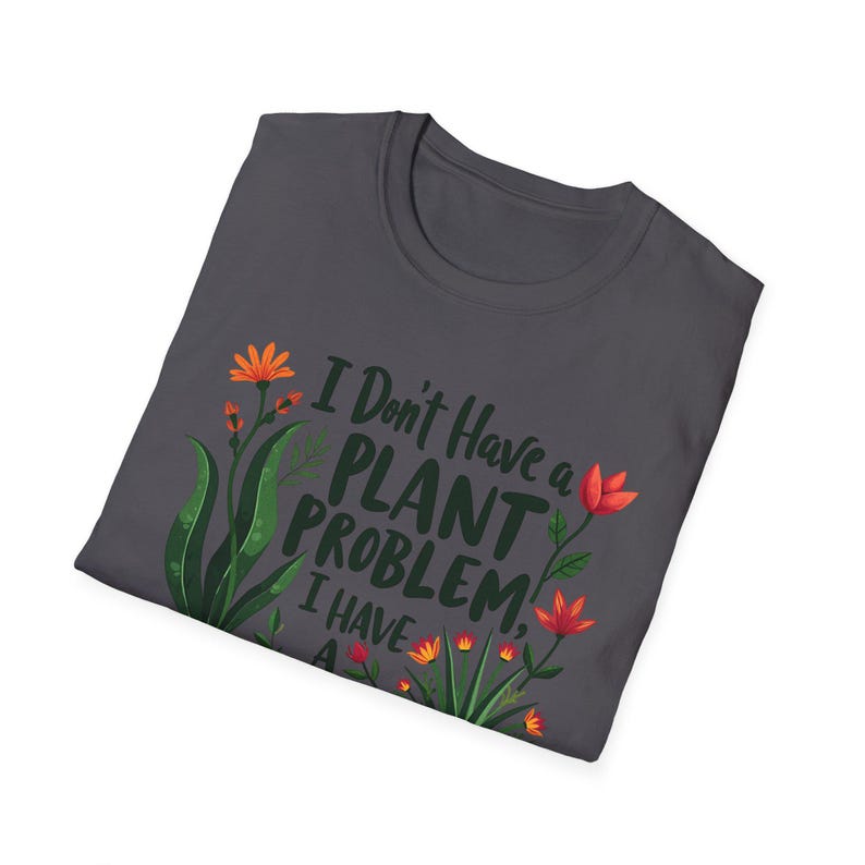 May include: Dark grey t-shirt featuring the text "I Don't Have a Plant Problem, I Have a..." and floral illustrations in orange and green. The shirt is folded.