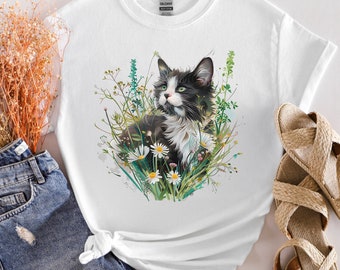 Könnte beinhalten: Weißes T-Shirt mit einer schwarz-weißen Katze, die in einem Feld mit Blumen sitzt. Die Katze hat grüne Augen und weiße Markierungen im Gesicht und auf der Brust. Die Blumen sind weiß und gelb.