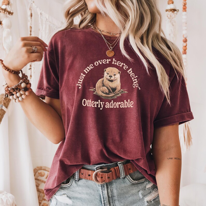 May include: A maroon t-shirt with a graphic of an otter and the text "Just me over here being Otterly adorable."