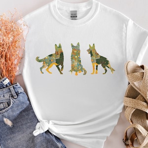 May include: White t-shirt with a graphic of three German Shepherd dogs. The dogs are outlined in a floral pattern with shades of green, yellow, and orange.