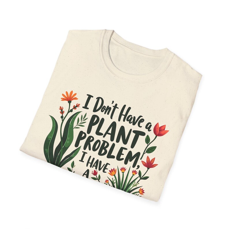 May include: Cream-coloured t-shirt featuring the text "I Don't Have a PLANT PROBLEM, I HAVE A..." with green leaves and orange and red flowers. The shirt is folded and has a round neckline.